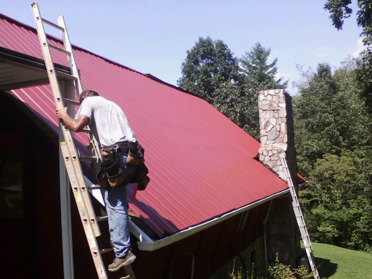 Roofer on a ladder beside a red metal roof and stone chimney — Soffit & Fascia Repair by Roofing Brothers in Simpson